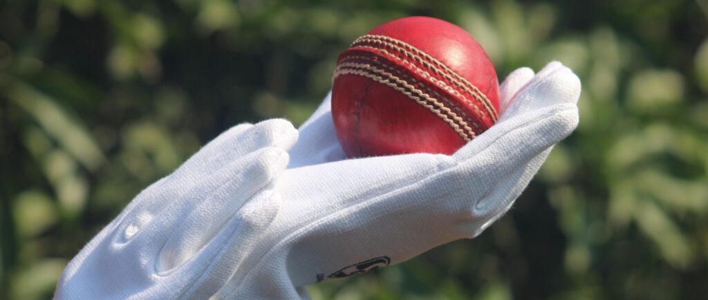 Detailed photo of a red cricket ball held by gloved hands outdoors.